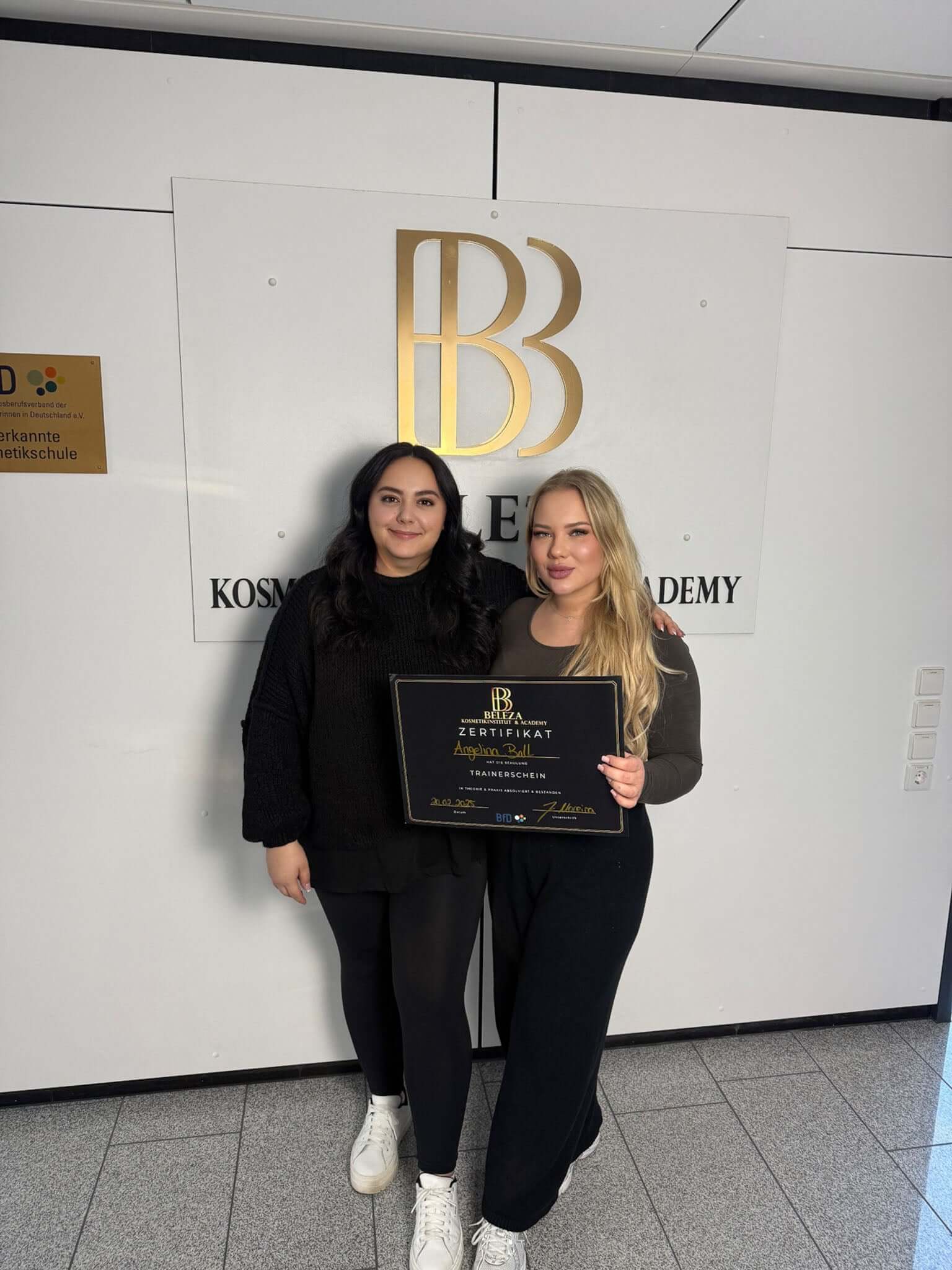 Two women proudly display a certificate in front of a modern academy logo, celebrating their achievement.