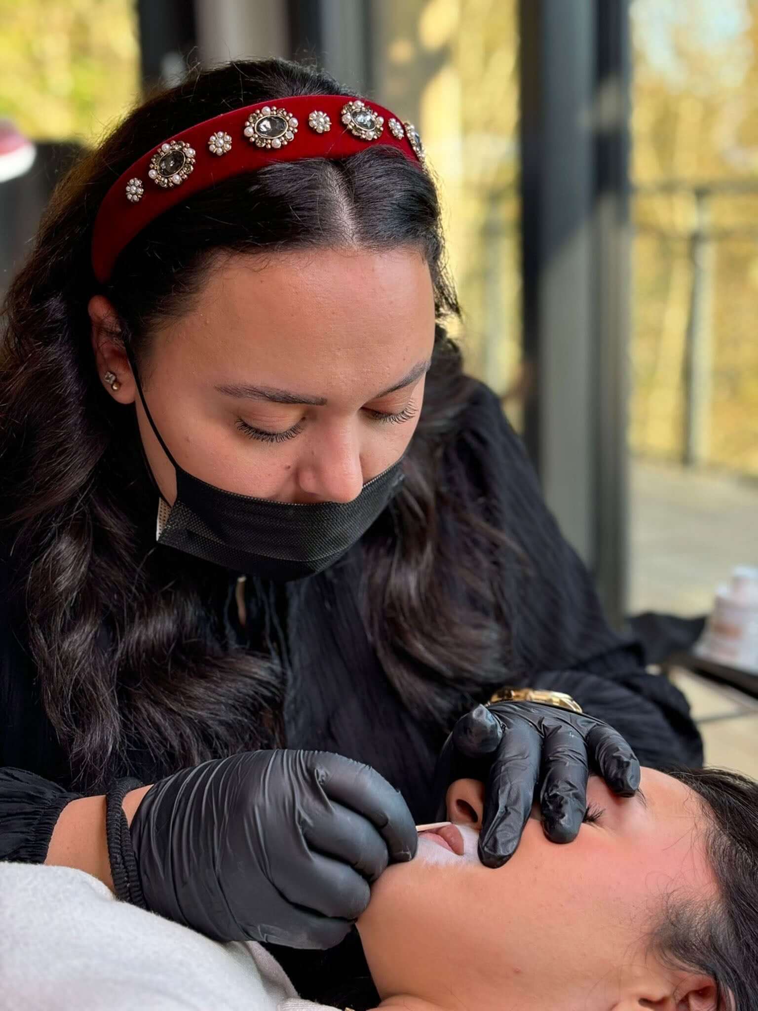 A beauty professional applying makeup with gloves on a client in a salon, wearing a stylish headband.