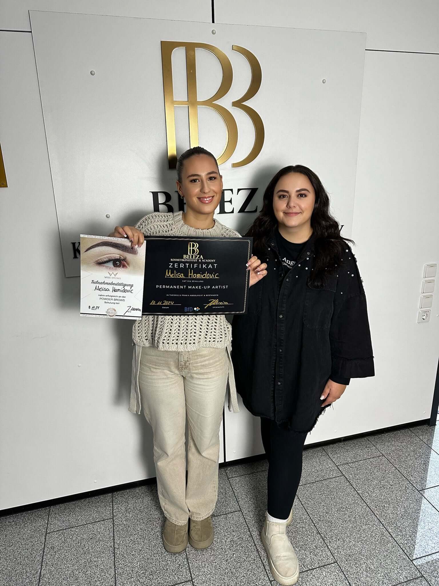 Two women holding a certificate and a diploma in a beauty studio, celebrating achievement together.