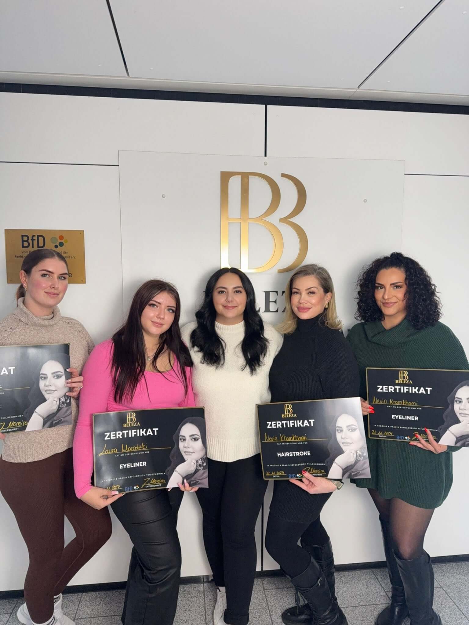 Five women celebrating certification achievements in beauty, holding certificates in front of a stylish backdrop.