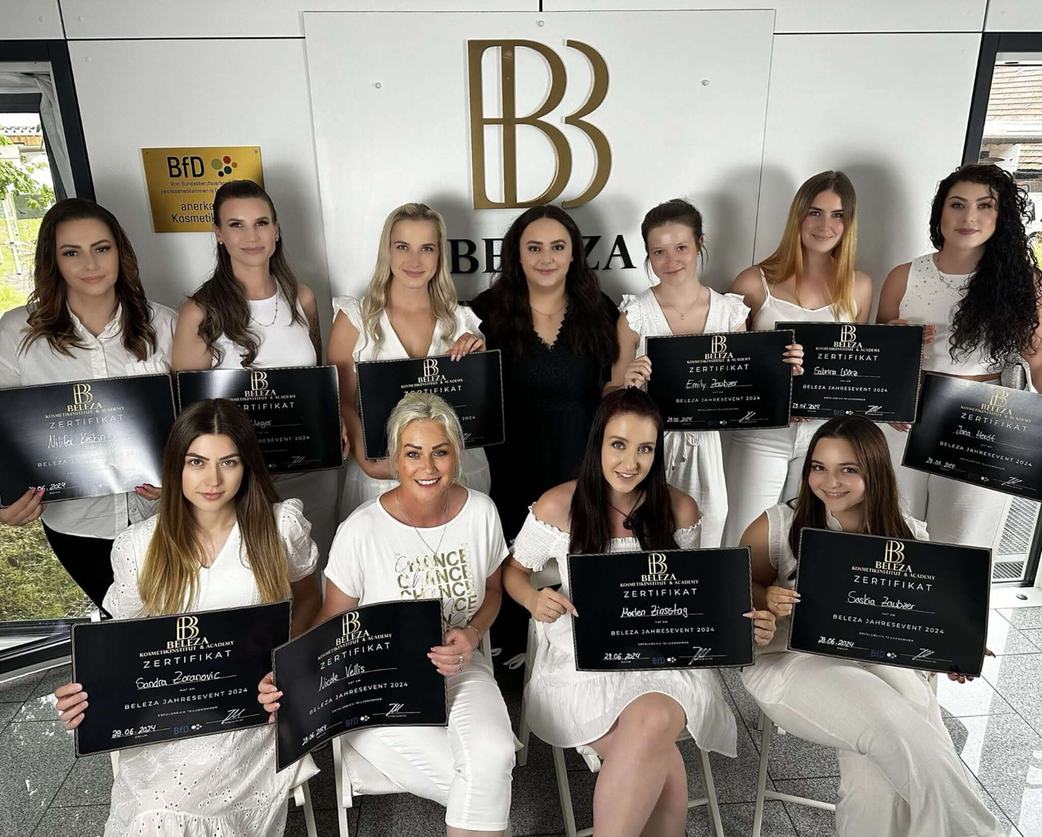 Group of graduates celebrating with certificates at a beauty school event, wearing white outfits.