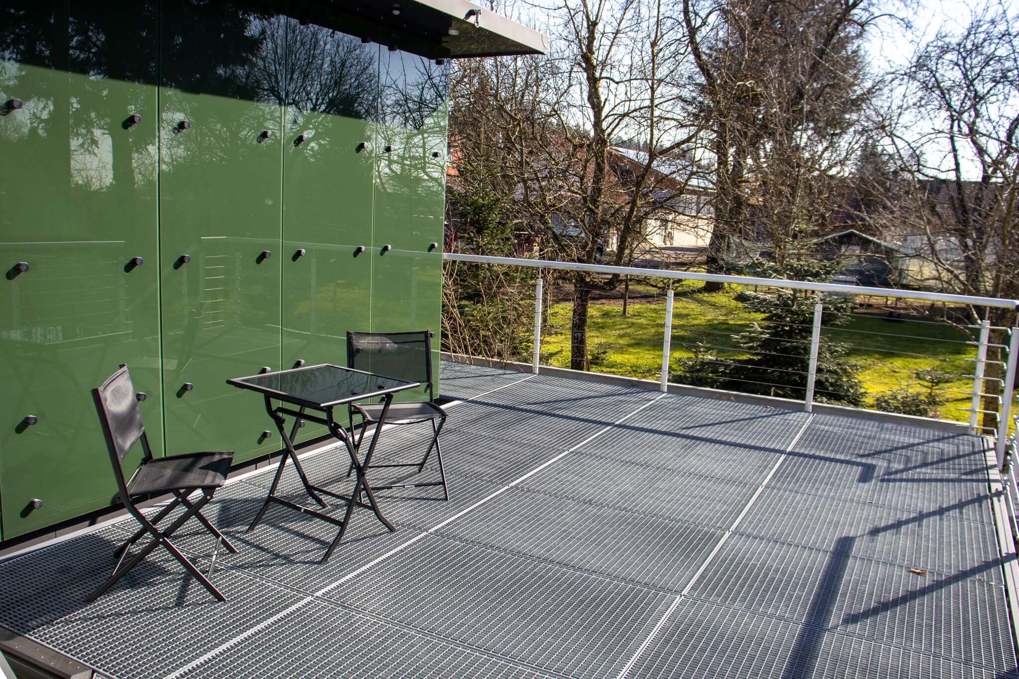 Modern outdoor balcony with a black table and chairs, surrounded by greenery in bright sunlight.