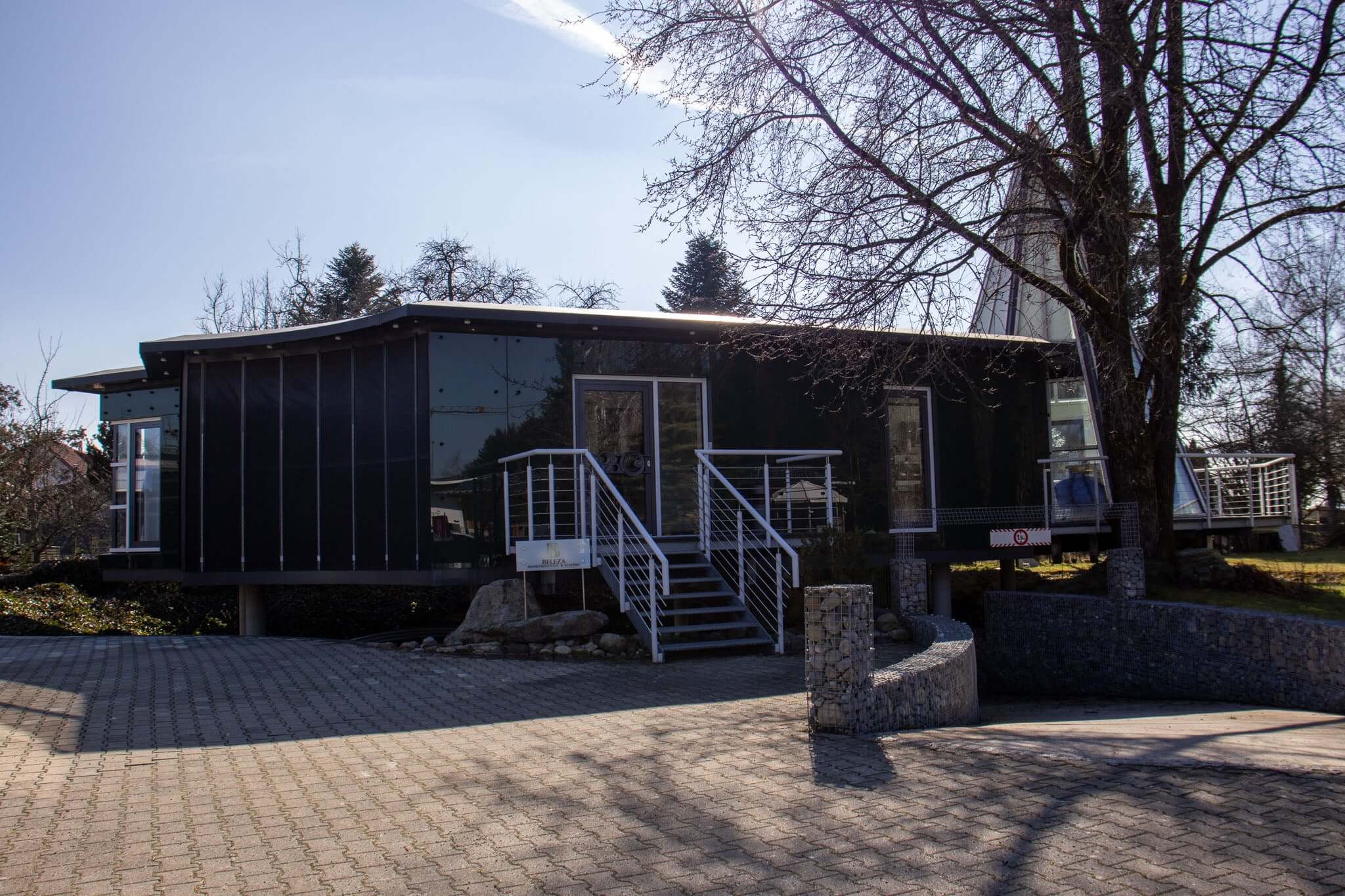 Modern black modular building with large windows and stairs on a sunny day, surrounded by trees.